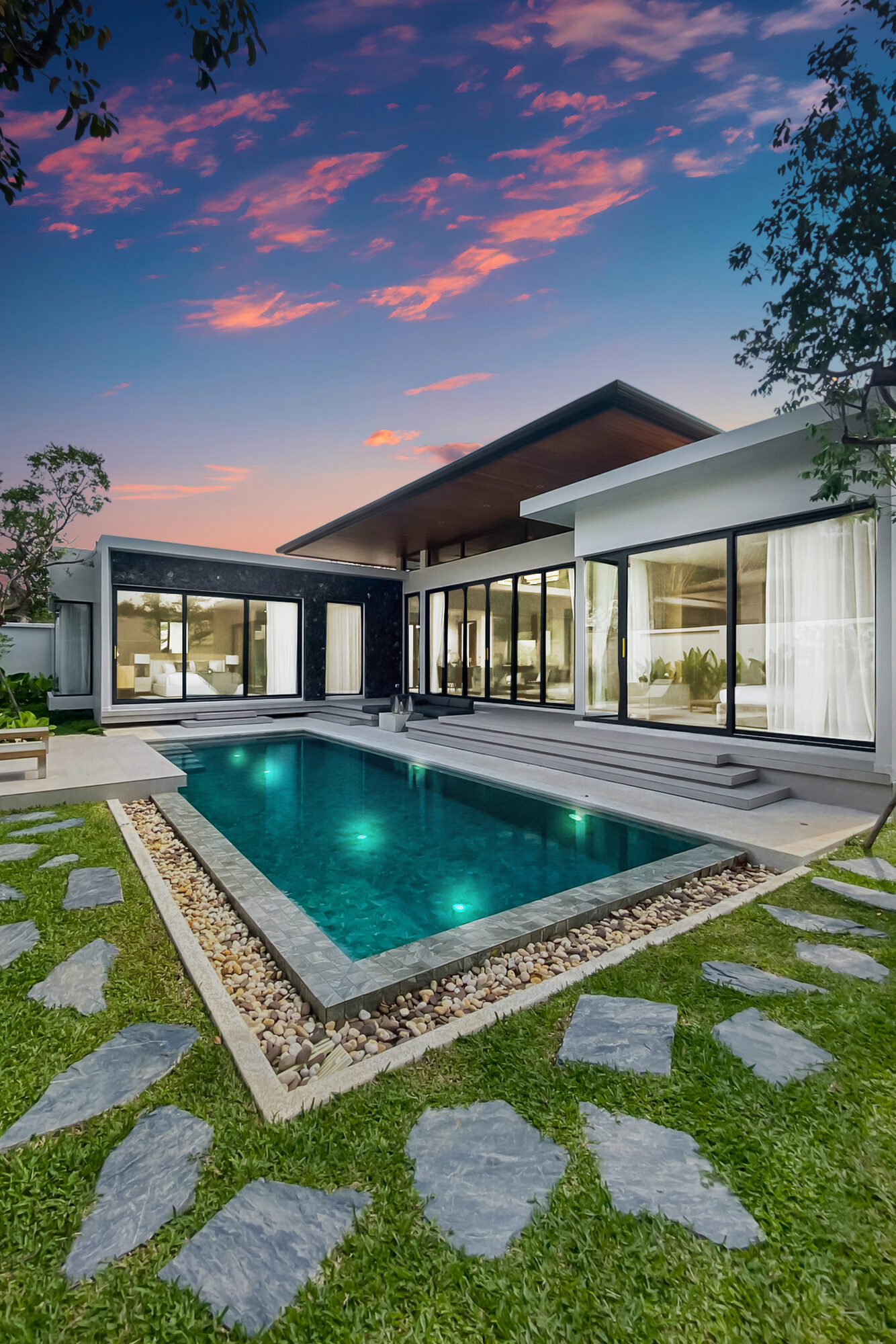 Villa Luma Botanica pool and villa at dusk, with stone stepping stones across the lawn and warmly lit interiors beyond